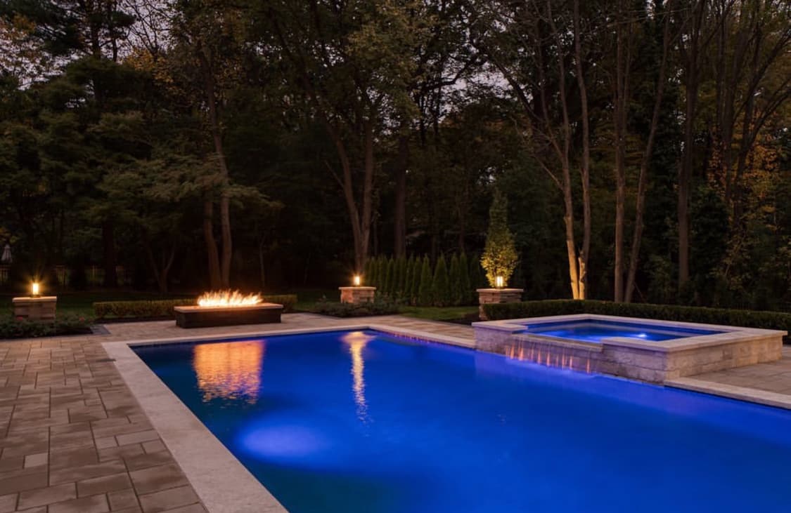 Pool with premium paver decking, fire bowl, raised spa, and landscape lighting at night.